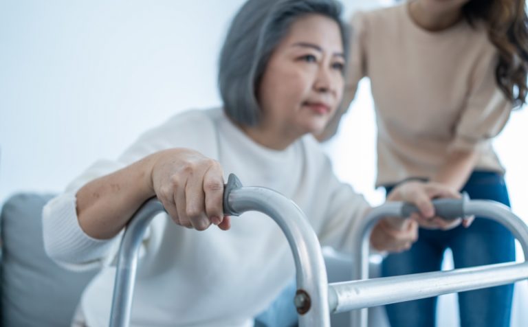 Asian young daughter support old disabled woman walk with walker at home. Beautiful girl help and take care of senior elderly mature handicap mom patient doing physical therapy in living room in house