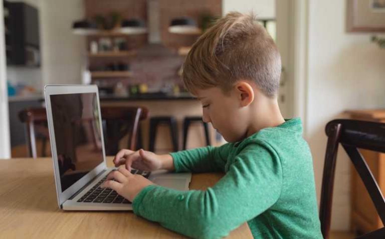 Side view of cute Caucasian boy using laptop at dining table in a comfortable home